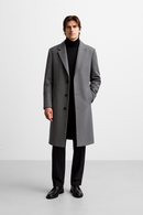 Um homem posa diante de fundo branco, usa o Casaco Sobretudo Masculino Longo Abaixo dos Joelhos da Zellum sobre suéter, calças e sapatos pretos, ressaltando conforto premium e o excelente acabamento da peça elegante.