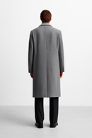 Um homem de costas veste o Casaco Sobretudo Masculino Longo Abaixo dos Joelhos da Zellum em tom cinza claro, com calça e sapatos pretos. O fundo branco valoriza o conforto premium e a sofisticação deste visual formal.