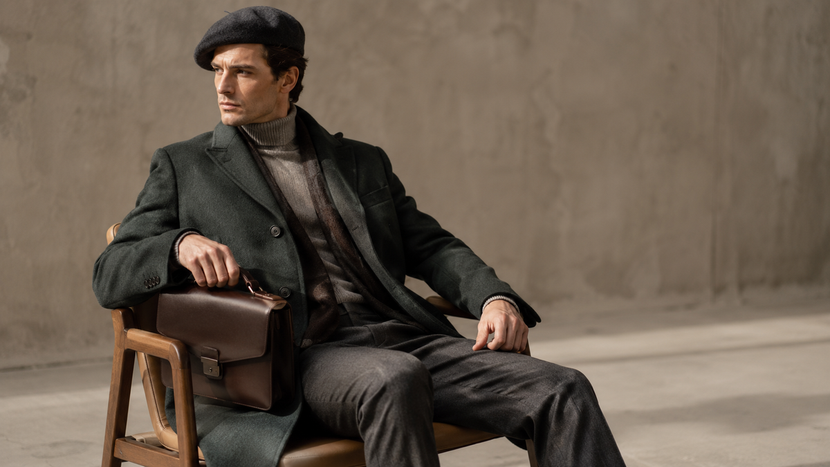 Homem com sobretudo longo verde escuro, boina cinza e pasta de couro, em look vintage sofisticado para o inverno