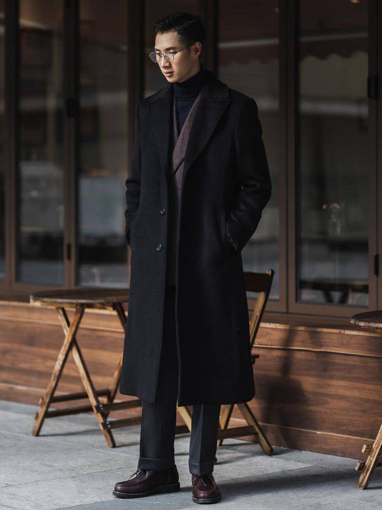 Homem usando sobretudo masculino preto elegante com cachecol vinho, calça de alfaiataria e sapatos sociais, em ambiente urbano sofisticado