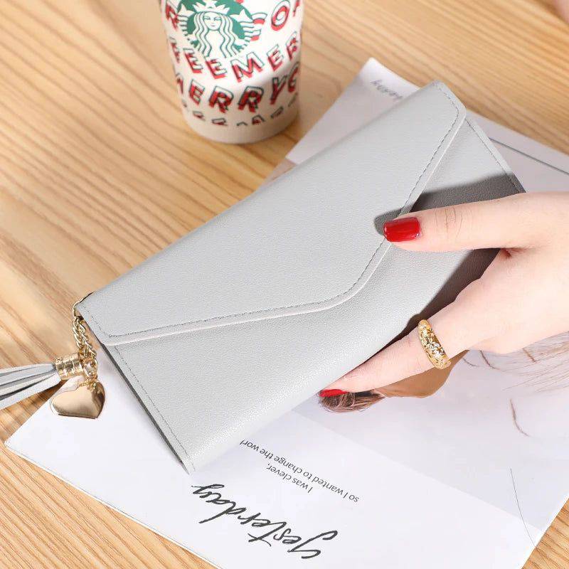 Mão feminina com anel dourado segurando carteira de couro multifuncional rosa claro com fecho de aba e borla, sobre um documento, com copo de café descartável ao fundo, em cenário casual.
