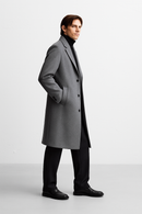 Um homem de perfil, contra um fundo branco, veste o Casaco Zellum Sobretudo Masculino Longo Abaixo dos Joelhos para conforto premium, combinado com calças pretas, um suéter de gola alta e sapatos sociais. Suas mãos repousam nos bolsos.