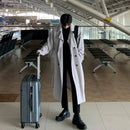 Homem em trench coat cinza longo, segurando mala de viagem em terminal de aeroporto