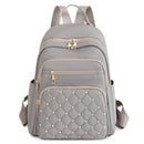Mochila feminina pequena de nylon cinza com bolso frontal acolchoado em matelassê com tachas douradas, múltiplos zíperes e alças ajustáveis.