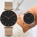 Dois relógios minimalistas com mostrador preto e pulseira mesh rosê, usados no pulso com pulseira delicada