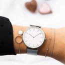 Relógio minimalista com mostrador branco e pulseira de aço mesh, combinado com pulseiras de corrente delicadas
