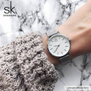 Relógio feminino SK com mostrador branco e pulseira em malha metálica, usado no pulso