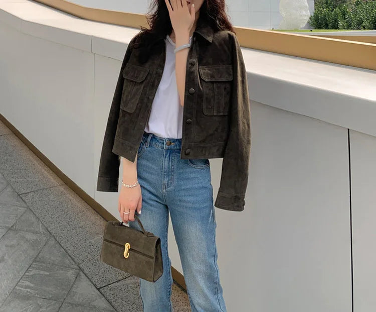 Ao ar livre, uma pessoa usa a Jaqueta Feminina de Pelo de Cabra Hircus da Zellum sobre uma camiseta branca e calça jeans azul, segurando uma bolsa marrom combinando. Seus cabelos soltos emolduram seu rosto enquanto ela está diante de uma parede curva moderna.