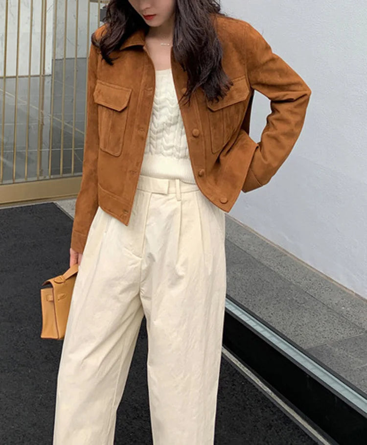 Uma mulher está ao ar livre, usando a Jaqueta Feminina de Pelo de Cabra Hircus da Zellum sobre uma blusa branca e calça creme. Ela segura uma clutch marrom e seus longos cabelos ondulados cobrem parcialmente seu rosto.