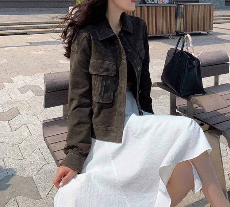 Uma mulher com longos cabelos escuros está sentada em um banco ao ar livre usando um Zellum Jaqueta Feminina de Pelo de Cabra Hircus com bolsos grandes sobre um vestido branco. Uma bolsa de mão preta repousa ao lado dela entre pedras de pavimentação e estruturas de madeira.