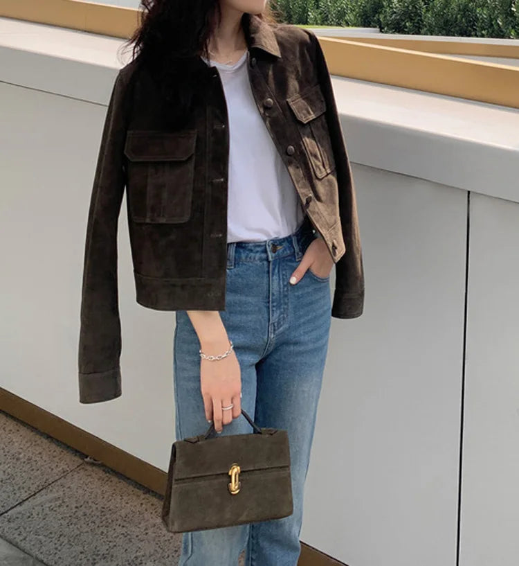 Uma mulher está ao ar livre usando um Zellum Jaqueta Feminina de Pelo de Cabra Hircus sobre uma camiseta branca e calça jeans azul, segurando uma bolsa marrom com fecho dourado. Sua mão direita está no bolso e seu rosto está parcialmente fora de quadro.