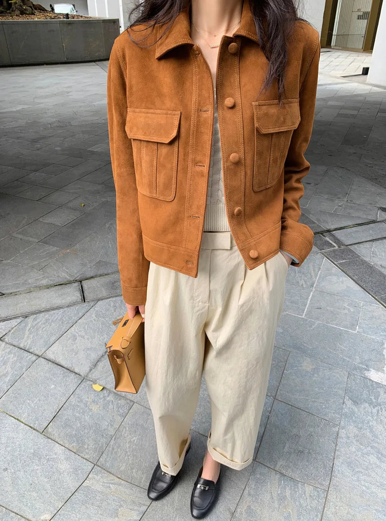 Uma pessoa usa a Zellum Jaqueta Feminina de Pelo de Cabra Hircus com bolsos e botões grandes, calça bege de cintura alta, blusa creme com nervuras, mocassins pretos e segura uma pequena bolsa marrom ao ar livre em meio à arquitetura moderna.
