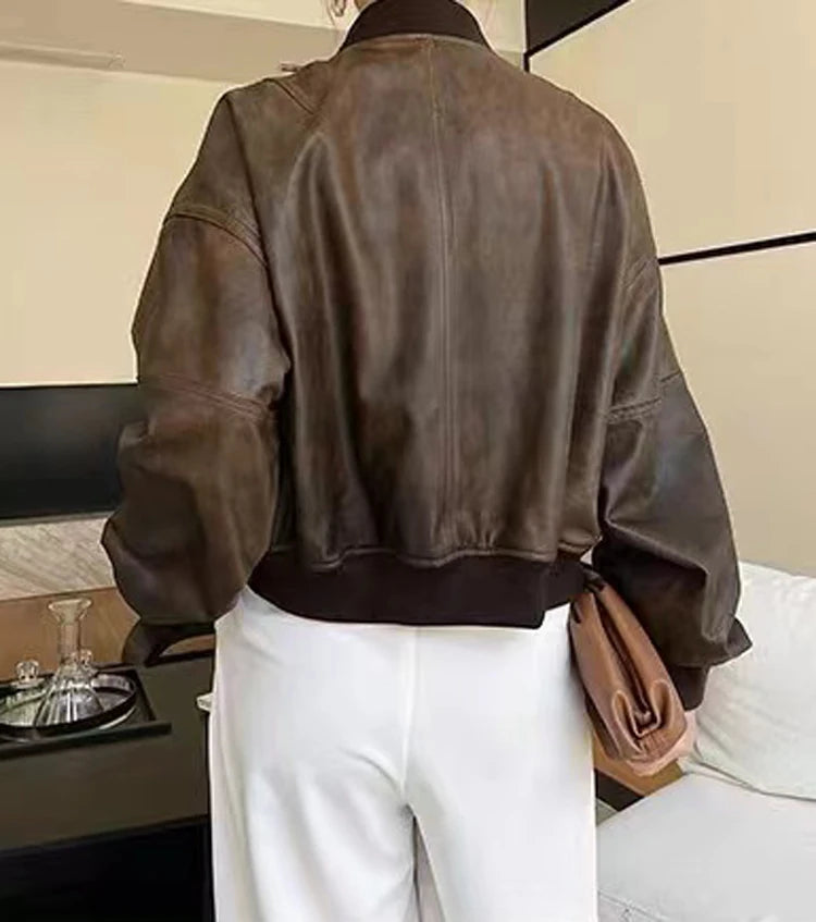 Uma mulher está em um ambiente interno, de costas para a câmera, usando uma Jaqueta Feminina de Couro Urban da Zellum e calças brancas de cintura alta. Ela segura uma bolsa marrom; um decantador de vidro e copos estão em uma mesa preta próxima.