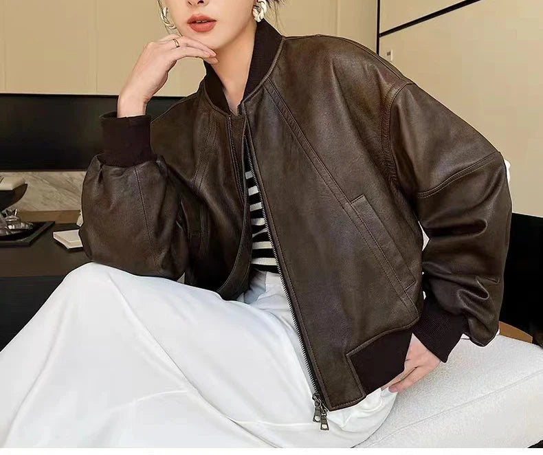 Uma pessoa se sente em uma cadeira branca vestindo a Jaqueta Feminina de Couro Urban da Zellum com punhos pretos, blusa listrada P&B e saia longa branca. Apoia o rosto na mão esquerda, mostrando brincos em um ambiente moderno neutro.