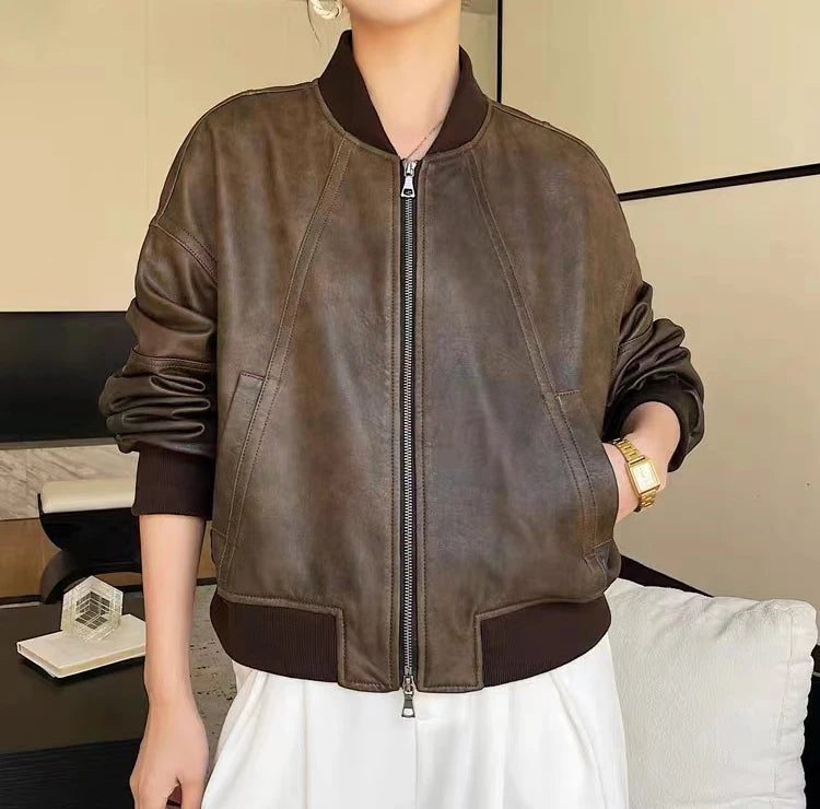 Uma pessoa em pé em um ambiente interno usando a Jaqueta Feminina de Couro Urban da Zellum, com punhos, gola e bainha canelados sobre uma roupa branca. Mãos nos bolsos com um relógio dourado. O quarto moderno mostra uma cama e uma mesa preta ao fundo.