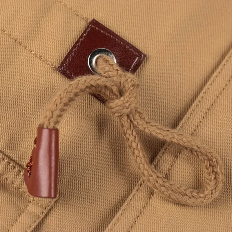 O close-up da lona bronzeada da Zellum Jaqueta Masculina Militar Forrada mostra um ilhó de metal reforçado com couro marrom com um cordão trançado preso por uma tampa de couro. As costuras pespontadas e o tecido texturizado destacam os detalhes robustos.