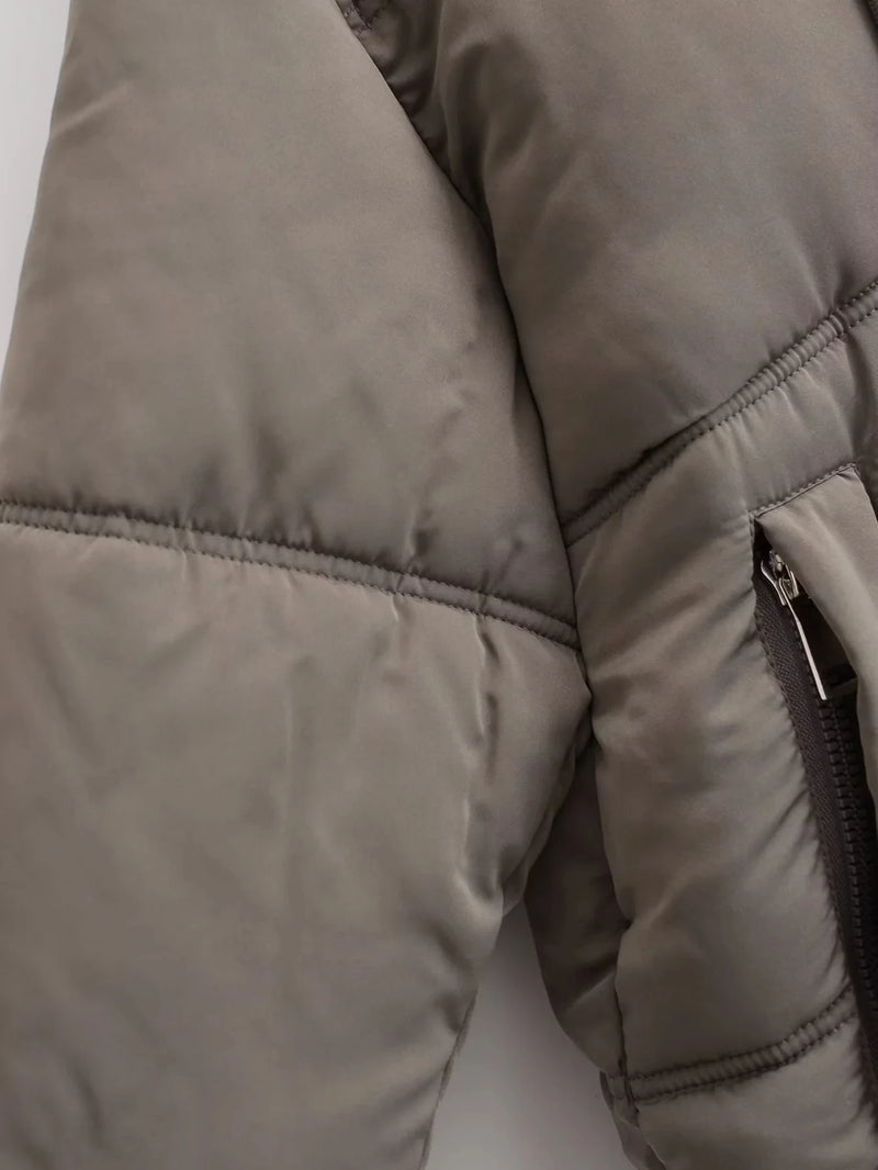 Close-up da Jaqueta Feminina Puffer para Inverno Zellum na cor taupe, com costuras horizontais, bolso com zíper e puxador de metal e tecido liso e levemente brilhante. A iluminação suave e difusa destaca parte da manga e do tronco.