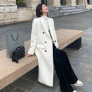 Mulher vestindo sobretudo longo off-white com abotoadura dupla, calça preta e tênis branco, sentada em banco de concreto