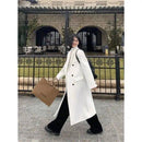 Mulher com sobretudo longo off-white, calça preta e bolsa bege em frente a edifício histórico