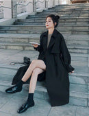 Mulher com trench coat preto longo abotoado duplo, bolsa acolchoada e sapatos pretos nos degraus.