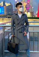 Homem em trench coat cinza longo com cinto, segurando bolsa, paisagem urbana ao fundo