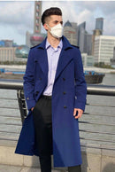 Homem com trench coat azul marinho abotoado duplo, camisa listrada e calça escura, em cenário urbano à beira de rio.
