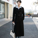 Homem usando trench coat preto longo com cinto sobre camisa listrada, segurando bolsa branca e preta em rua urbana