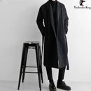 Homem em sobretudo longo preto com cinto e gola aberta, calça preta e sapatos pretos.