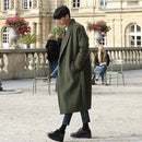Homem usando sobretudo longo verde militar com cinto, jeans escuro e botas pretas em jardim clássico europeu