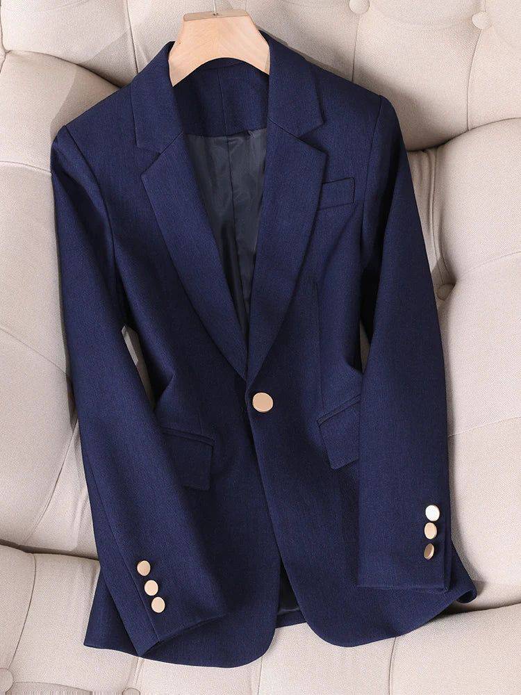 Blazer feminino alfaiataria azul marinho com botão dourado, apoiado em sofá claro