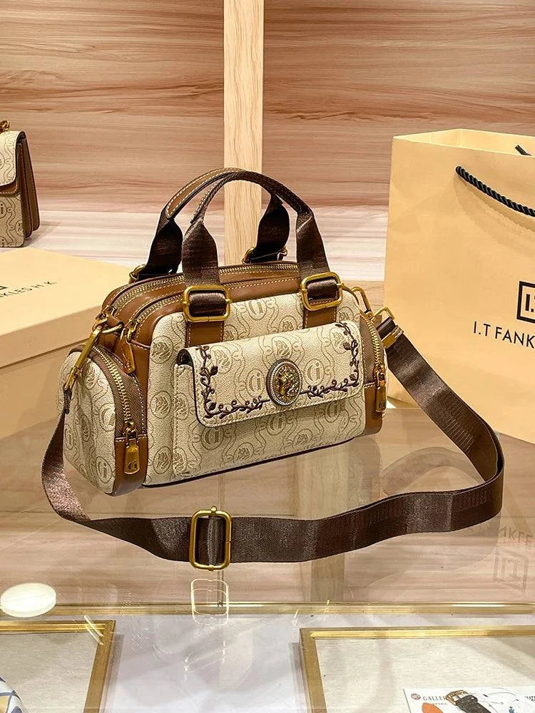 Bolsa Baú Feminina marrom e bege, tecido estampado. Bolso frontal bordado com leão dourado, zíperes laterais. Alça transversal ajustável. Elegante e estruturada.