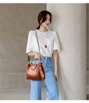 Mulher com blusa branca de renda, calça jeans clara e bolsa de couro marrom com alça de metal no ombro