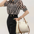Bolsa de couro creme com alças duplas marrom, segurada por mulher com blusa listrada preta e branca
