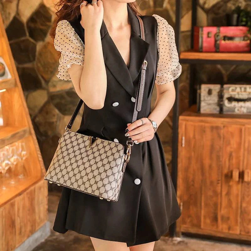 Bolsa Feminina de Couro Elite bege, estampa de monograma, logo "V" dourado. Alça de corrente e transversal. Usada por modelo em vestido. Sofisticada e elegante.
