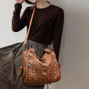 Bolsa de couro marrom com detalhes trançados, alças duplas e alça longa, em look com blusa preta e saia plissada