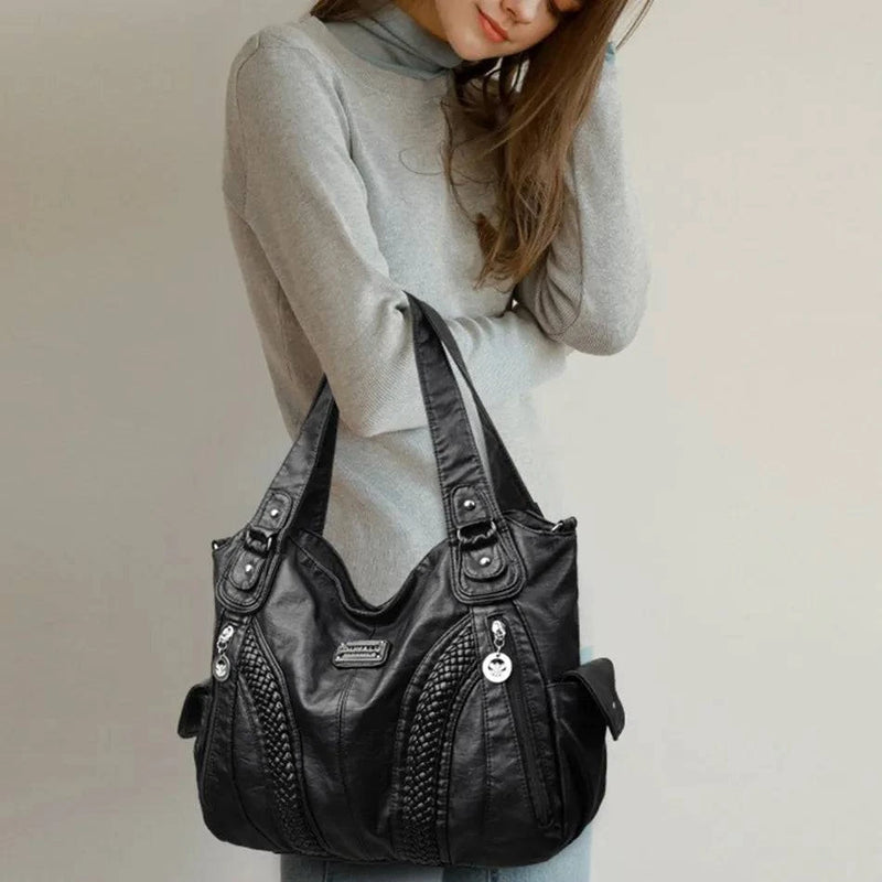 Bolsa feminina de couro genuíno preta, modelo Charmonia, com detalhes trançados, alças duplas, zíper e detalhes prateados, usada com blusa de gola alta cinza