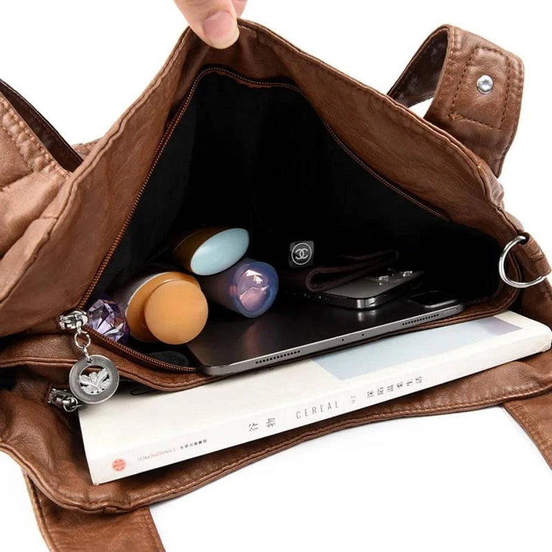Bolsa feminina de couro legítimo marrom modelo Charmonia, aberta e exibindo seu interior espaçoso com cosméticos, celular, tablet e livro organizados, mostrando sua capacidade de armazenamento.