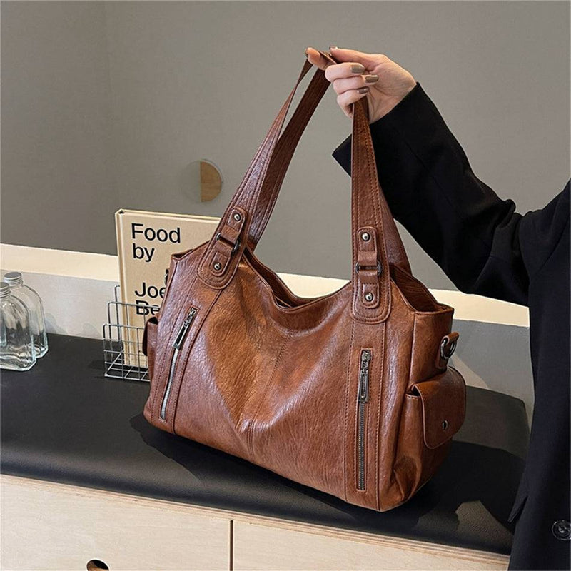 Mulher segurando bolsa feminina hobo em couro marrom com alças reforçadas, zíperes frontais e bolso lateral com botão.
