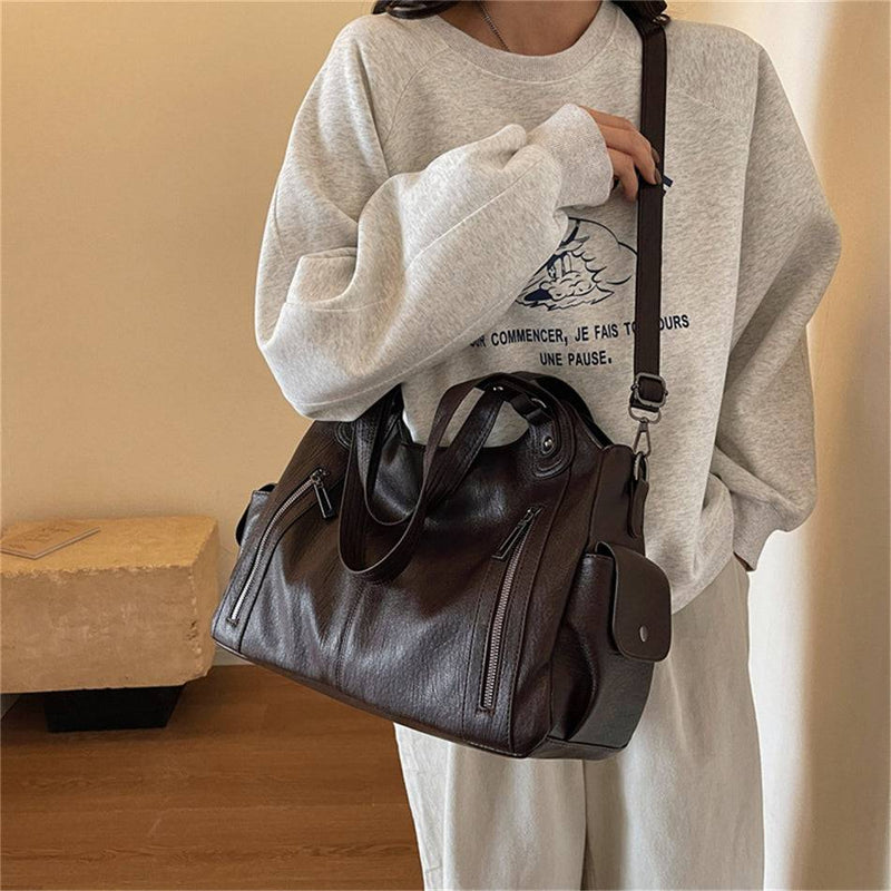 Mulher segurando bolsa feminina hobo em couro marrom escuro com alça transversal ajustável, bolsos laterais e zíperes frontais.