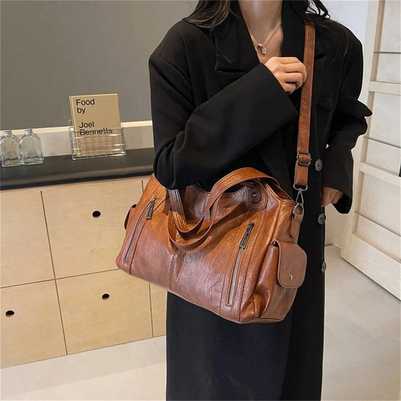 Mulher segurando bolsa feminina hobo em couro marrom com alça transversal, zíperes frontais e bolso lateral com fecho.