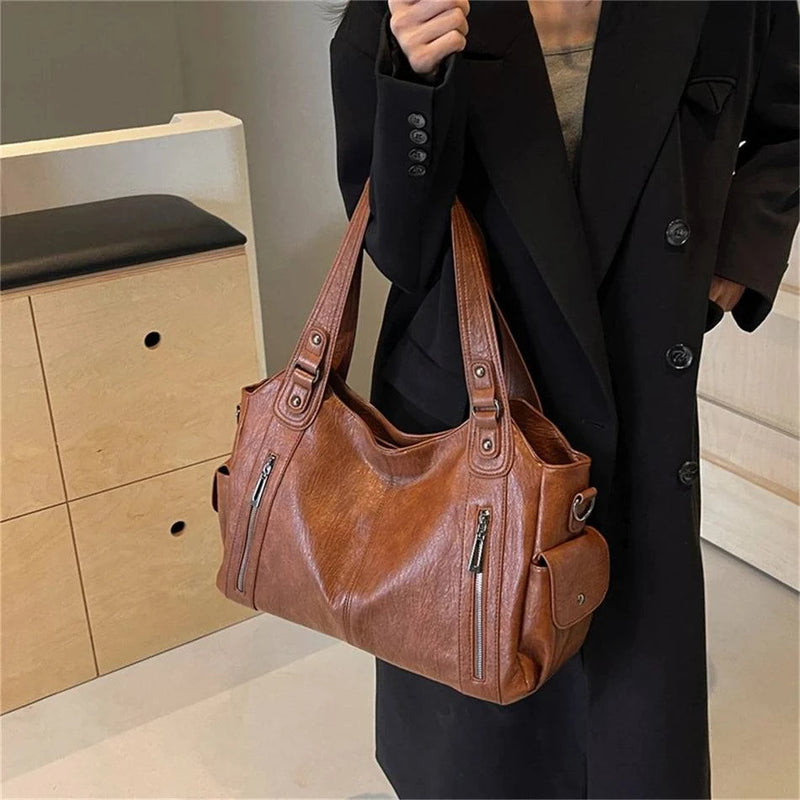 Mulher segurando bolsa feminina hobo em couro marrom com detalhes de zíper frontal, bolsos laterais e alças reforçadas.