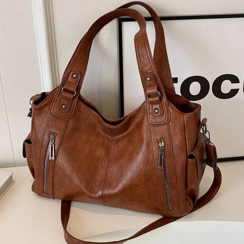 Bolsa feminina hobo em couro marrom com alças duplas, bolsos laterais e detalhes em zíper metálico frontal.