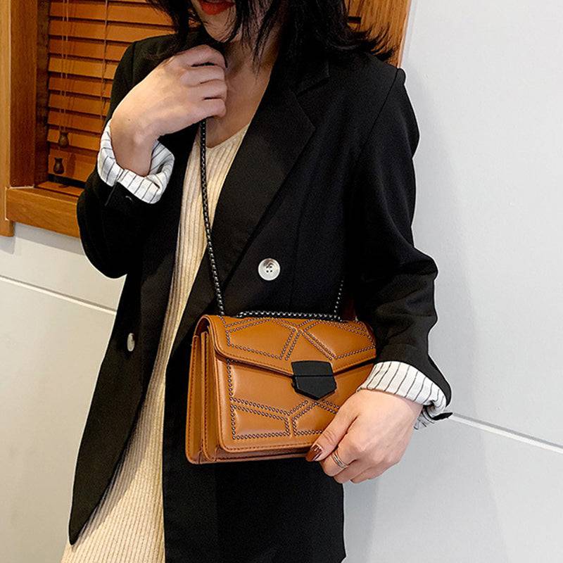 Bolsa Feminina Tiracolo Estruturada na cor caramelo com design matelassê de rebites e alça de corrente escura, combinando com um blazer preto, ideal para um visual elegante e sofisticado.