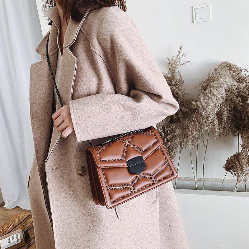 Bolsa Feminina Tiracolo Estruturada caramelo com design matelassê de rebites, fecho geométrico preto e alça de corrente escura, combinando perfeitamente com um casaco bege, ideal para looks de outono/inverno.