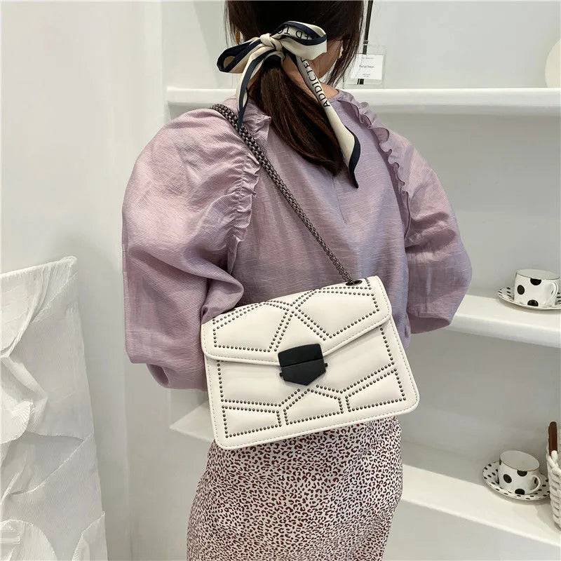 Bolsa Feminina Tiracolo Estruturada na cor creme com design matelassê de rebites e alça de corrente escura, exibida nas costas em um look casual, destacando o caimento da peça.