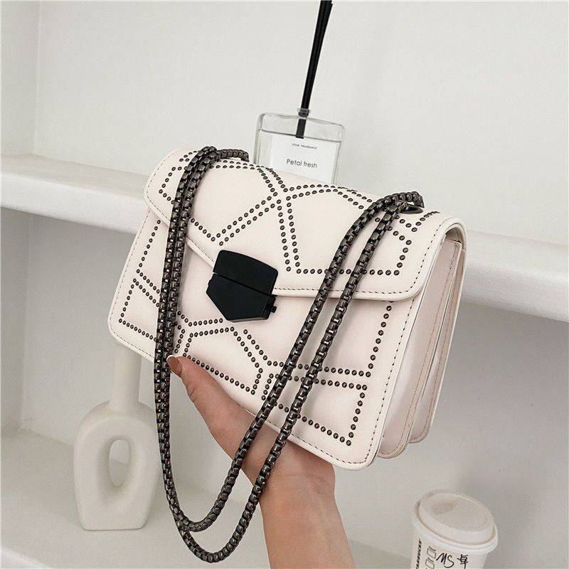Bolsa Feminina Tiracolo Estruturada na cor creme, com design matelassê de rebites, fecho frontal geométrico preto e alça de corrente escura, destacando os detalhes modernos.