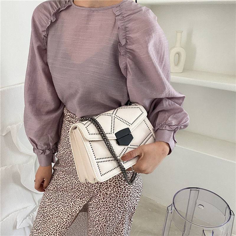Bolsa Feminina Tiracolo Estruturada na cor creme com design matelassê de rebites, fecho geométrico preto e alça de corrente escura, exibida na frente de um look casual, destacando seu estilo e versatilidade.