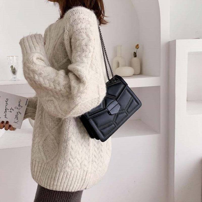 Bolsa Feminina Tiracolo Estruturada preta com design matelassê de rebites e alça de corrente, exibida lateralmente, mostrando a forma estruturada e o fecho frontal geométrico.