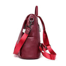 Perfil lateral da bolsa mochila antifurto de couro vinho, com alças de tecido vermelho, mosquetão prateado e zíper lateral visível, destacando a profundidade da bolsa.