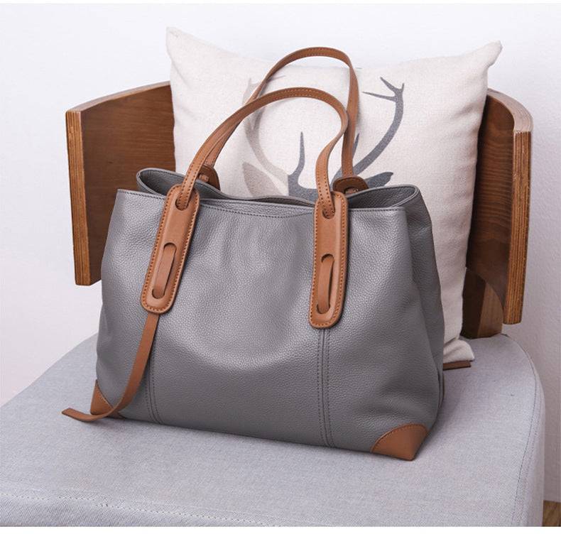 Bolsa de ombro feminina em couro legítimo, cor cinza com alças em marrom, sobre cadeira de madeira clara.
